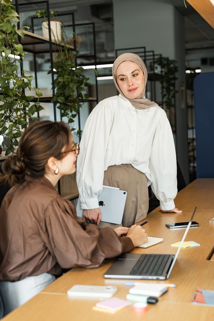 Two businesswomen in a modern office engaged in a work discussion, using laptops and notes.